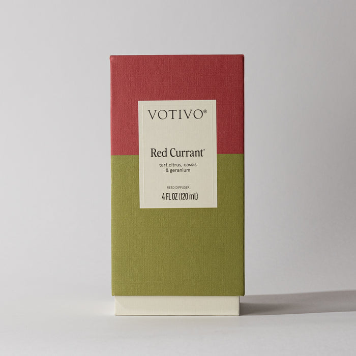 A Votivo Red Currant Reed Diffuser (4 fl oz/120 ml) with a red and olive green box design, fiber reeds, and a label describing tart citrus, cassis & geranium notes stands on a white surface.