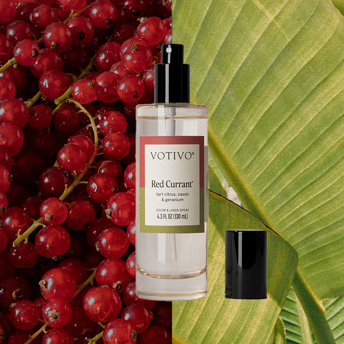 A clear Votivo Red Currant Room & Linen Spray bottle with a black cap sits against red currant berries and a green leaf, emphasizing the spray’s fresh, fruity scent.