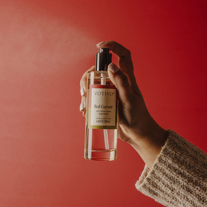 A hand in a beige sweater holds Votivo Red Currant Room & Linen Spray, misting its fruity scent against a solid red background.