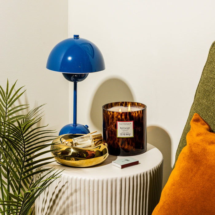 A blue lamp, Votivos Red Currant Tortoise Candle, a gold tray with matches, and a green sofa with an orange cushion rest on a white table by a small plant against a white wall.
