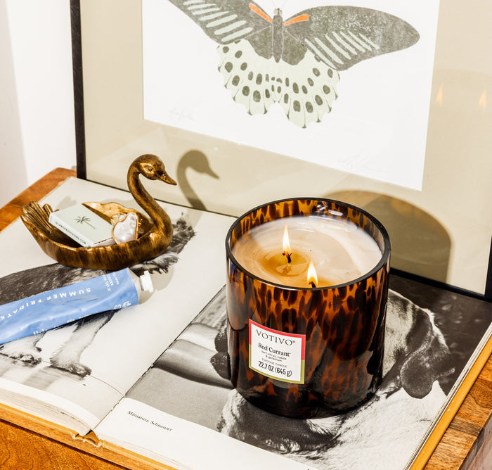 A glowing Votivo Red Currant Tortoise Candle sits in tortoiseshell glass atop an open book, beside a brass swan dish, a blue tube, and a butterfly illustration in the background.