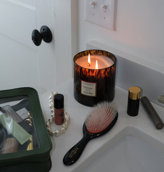 A Votivo Red Currant Tortoise Candle sits lit on the bathroom counter beside a glass makeup bag, lip gloss, pearl bracelet, hairbrush, and makeup products near a white sink and door.