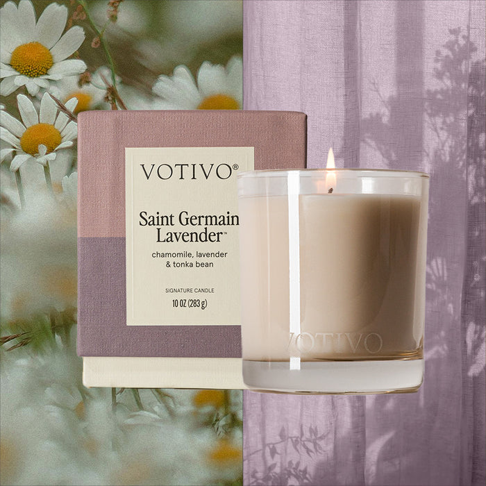 A lit Votivo Saint Germain Lavender 10 oz. Signature Candle sits in a clear glass by its box, surrounded by chamomile flowers and sheer lavender fabric. This soy blend candle features notes of chamomile, lavender, and tonka bean.