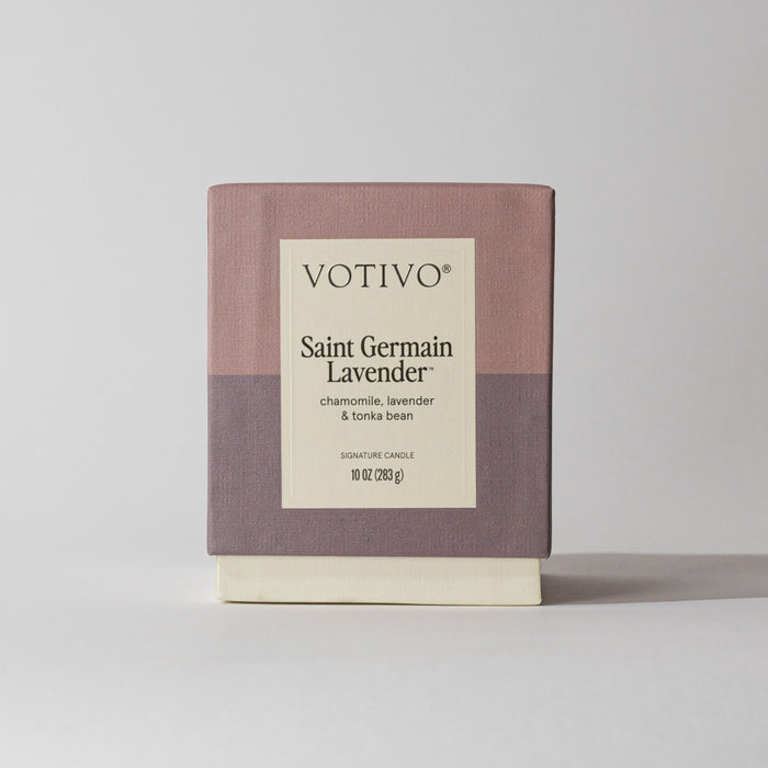 A Votivo Saint Germain Lavender 10 oz. Signature Candle with chamomile, lavender, and tonka bean in a minimalist mauve and lavender box stands out against a plain background. This soy blend wax candle is the perfect relaxing accent.