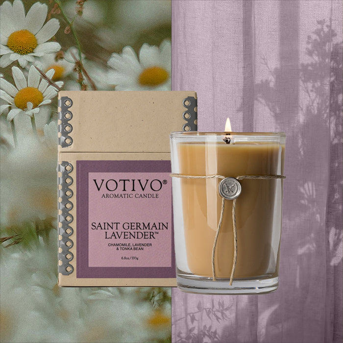 The Votivo Saint Germain Lavender 6.8 oz. Aromatic Candle, made with a soy blend and in a glass holder, sits lit beside its box amid daisies and purple fabric, decorated with twine and a round silver charm.