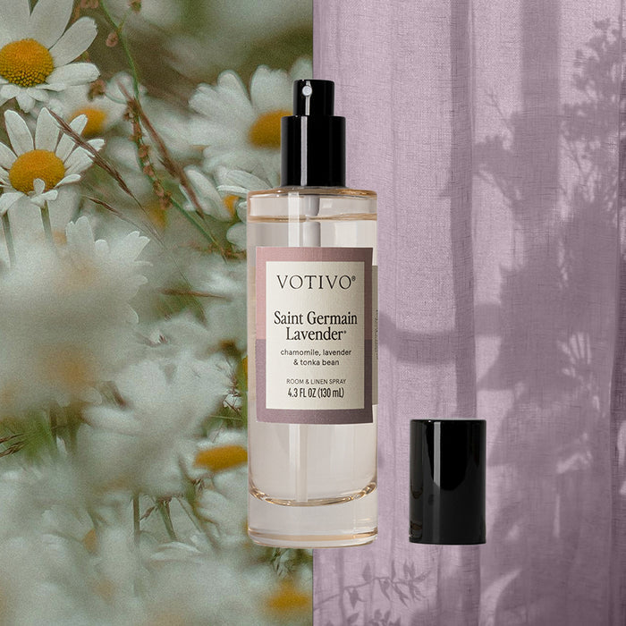 A clear spray bottle labeled Votivo Saint Germain Lavender Room & Linen Spray stands upright with its black cap beside it, set against chamomile flowers and a lavender curtain to highlight its soothing lavender fragrance.