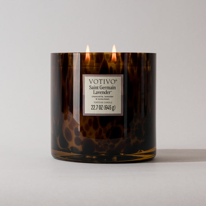 A Votivo Saint Germain Lavender Tortoise Candle with two lit wicks sits on a plain surface. This 22.7 oz (645 g) candle features a brown, speckled design and is made with a soy blend wax.