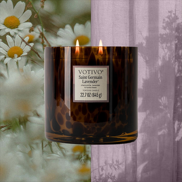 The Votivo Saint Germain Lavender Tortoise Candle, made from a soy blend wax with two flames in brown glass, burns in front of white daisies and a sheer lavender curtain.