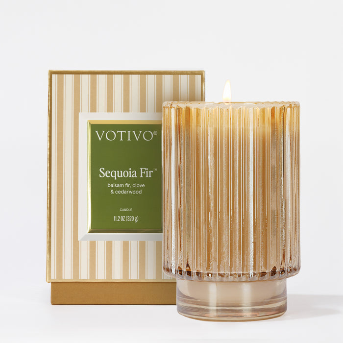 The Votivo Sequoia Fir Holiday Candle features a soy blend wax in a ribbed glass jar with a green label. Notes of balsam fir, clove, and cedarwood fill your space as it glows beside its festive striped box.