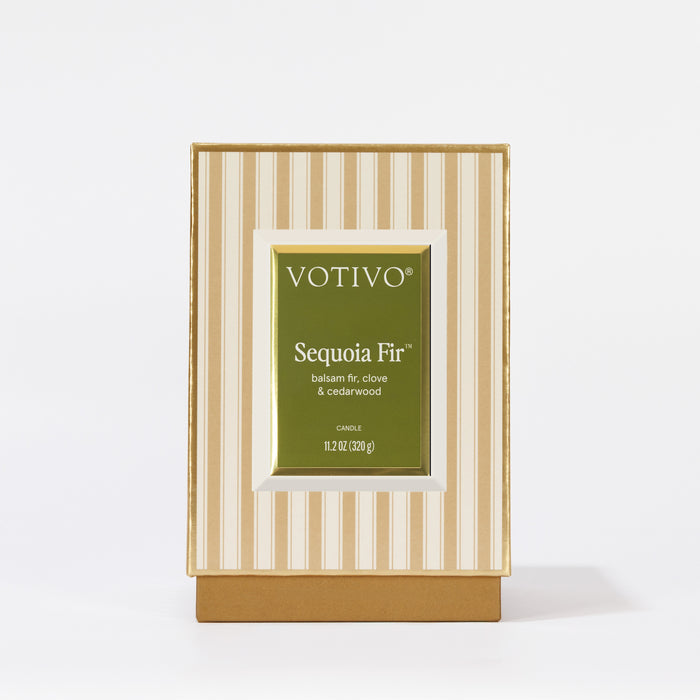 A Votivo Sequoia Fir Holiday Candle, crafted with premium soy blend wax, stands upright on a white surface in its box featuring a green label with white text and a striped beige and cream background.