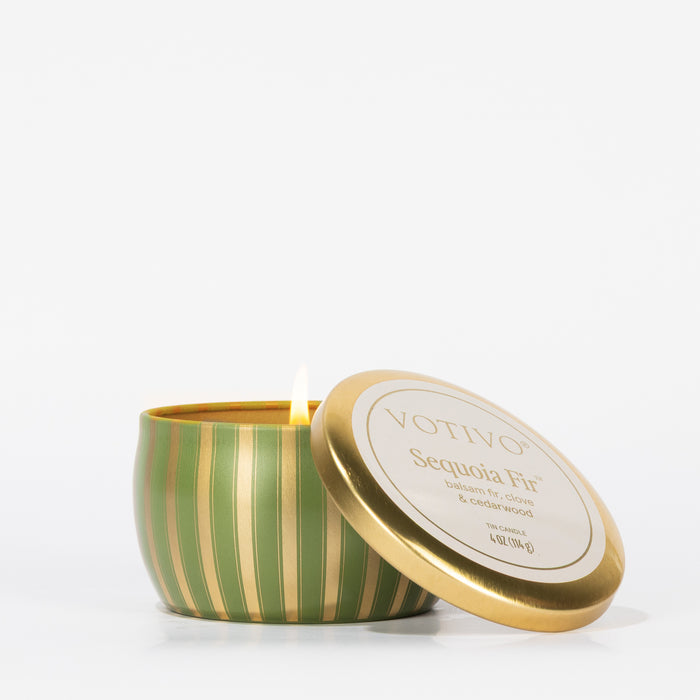 A lit Votivo Sequoia Fir Holiday Tin Candle in a green and gold striped tin, lid set aside, sits against a plain white background. The label displays the brand and scent name.