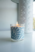 A lit candle in a glass holder with a blue coastal pattern sits on a white surface. A textured white vase is in the background.
