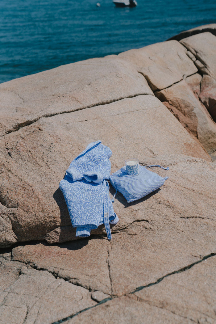 Baby blue patterned cotton robe, matching bag, and candle lie on sunlit rocks by the sea, with a boat visible in the background.