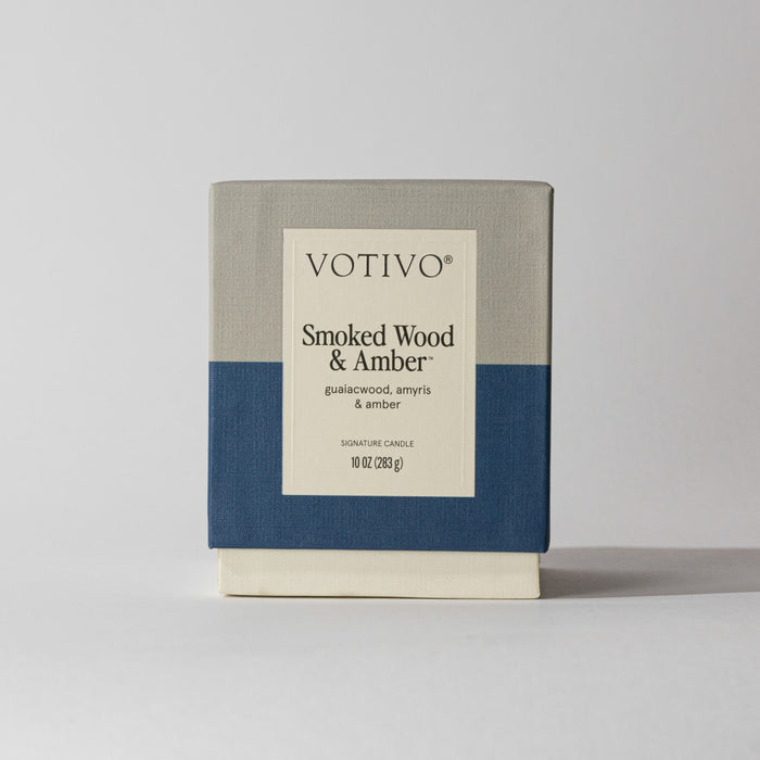 The Votivo Smoked Wood & Amber 10 oz. Signature Candle features guaiacwood, amyris, and amber notes. Made with soy blend wax, its box displays a beige, gray, and navy color block design on a simple light background.