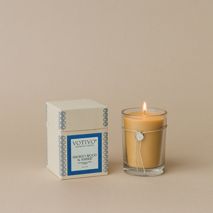 A lit beige Votivo Smoked Wood & Amber 6.8 oz. Aromatic Candle in a clear glass holder sits beside its box, made with soy blend wax, against a plain beige background.