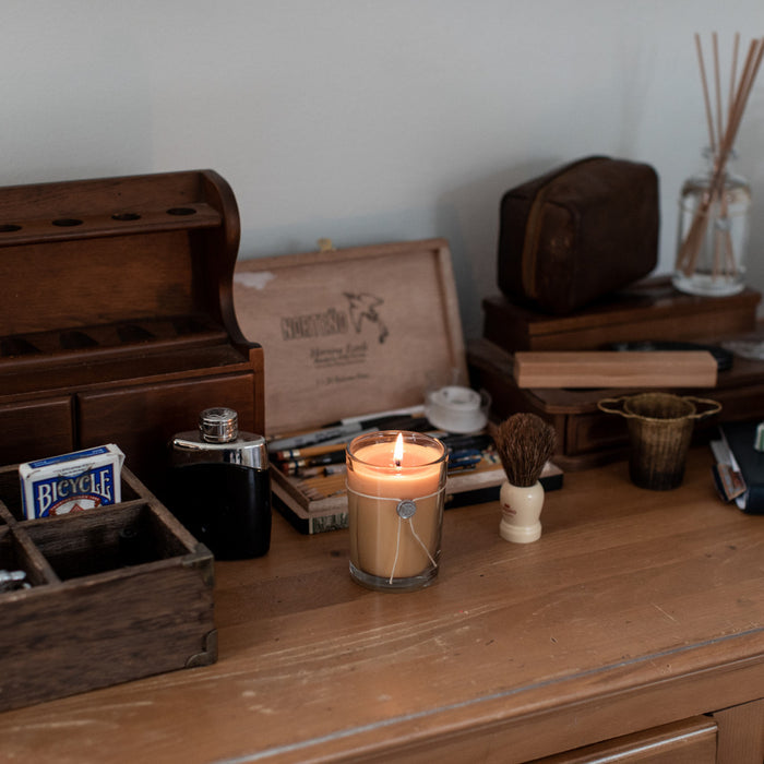 The Votivo Smoked Wood & Amber 6.8 oz. Aromatic Candle rests on a wooden desk, surrounded by a playing card deck, shaving brush, metal flask, wooden box, grooming tools, and small containers.