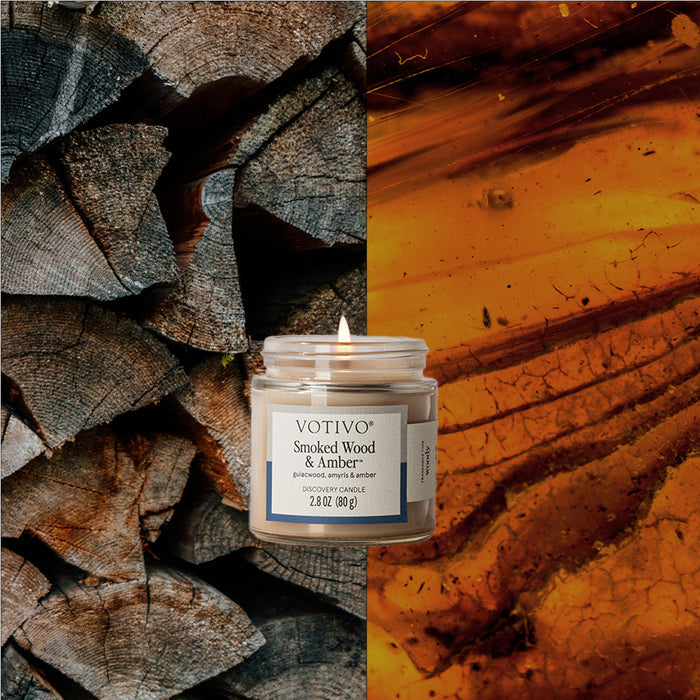 A lit Votivo Smoked Wood & Amber Discovery Candle, made with a soy blend wax, sits between cut firewood and amber resin, showcasing its signature fragrance notes.