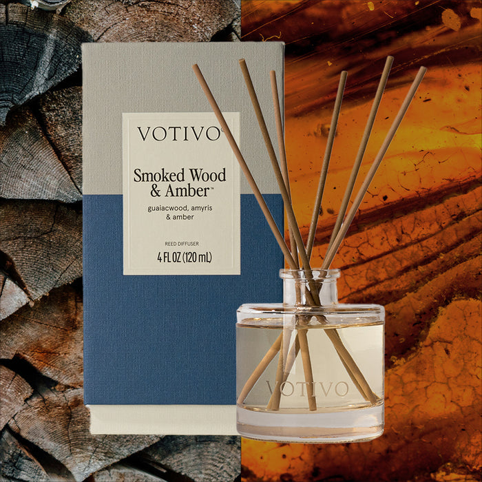 Votivo’s Smoked Wood & Amber Reed Diffuser features fiber reeds in a clear glass bottle, presented in a blue and beige box, displayed with wood logs and glowing amber.