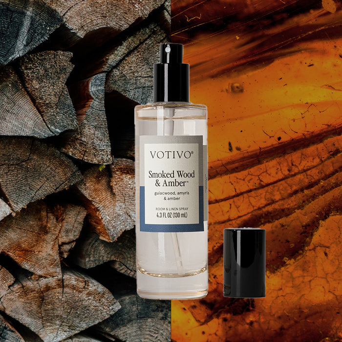 A bottle of Votivo Smoked Wood & Amber Room & Linen Spray sits beside stacked firewood and amber resin, its black cap nearby—an inviting way to refresh any space.