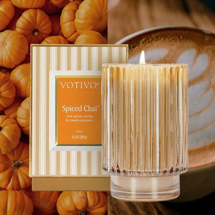 A Votivo Spiced Chai Holiday Candle made with soy blend wax is displayed in a ribbed glass holder with its striped box, surrounded by small pumpkins and a leaf-art latte in the background.