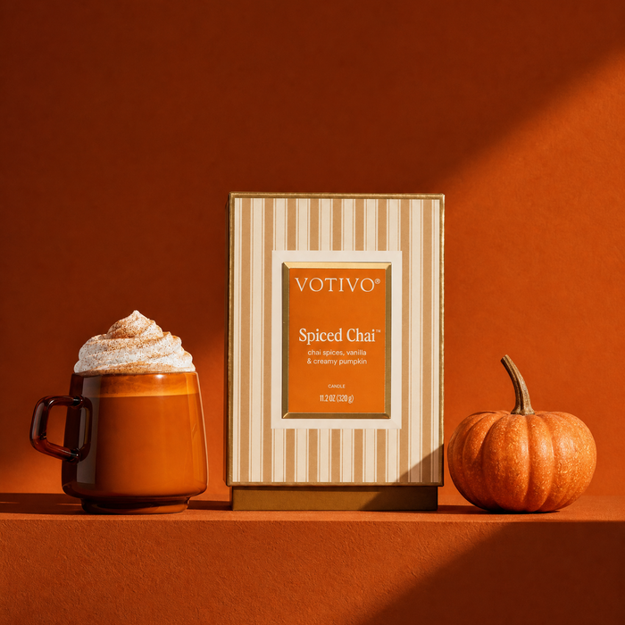 Votivo Spiced Chai candle 
box standing on a warm terracotta surface, flanked by a whipped-cream-topped mug and a small pumpkin, in a cohesive autumn color palette.