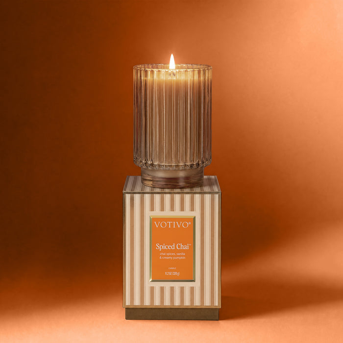 A lit Votivo Spiced Chai Holiday Candle in a ribbed glass jar rests on its striped box against an orange backdrop. Made with soy blend wax, it features notes of cinnamon, vanilla, and creamy musk.