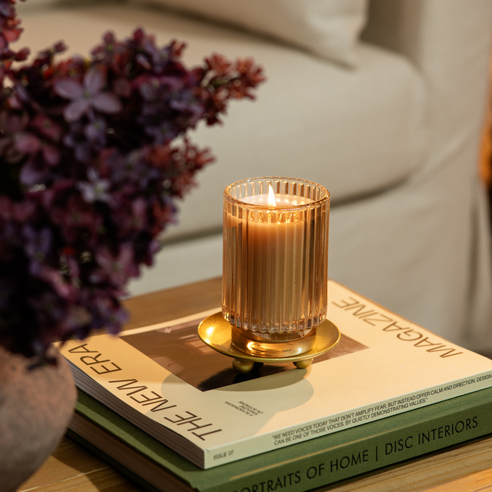A lit candle in a ribbed clear glass holder with a gold-toned base, styled on top of books beside a blurred purple floral arrangement in a softly lit indoor setting.