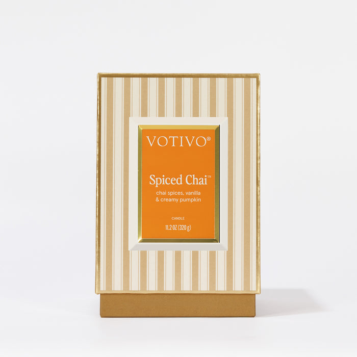 The Spiced Chai Holiday Candle by Votivo features cream and gold stripes with an orange label. Enjoy chai spices, vanilla, and creamy pumpkin in a premium soy blend wax for a cozy seasonal aroma.