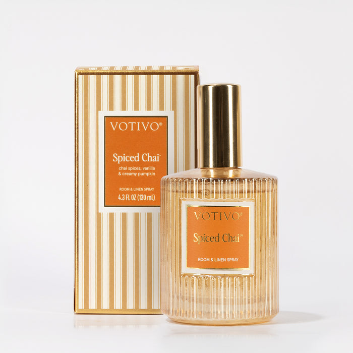 A Votivo Spiced Chai Room & Linen Spray with a gold cap is displayed before its matching striped box. The orange label features white text, and the spray is fabric safe for versatile holiday freshness.