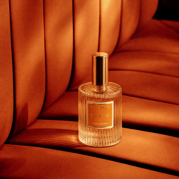 A ribbed clear glass Spiced Chai room spray bottle with a gold cap sitting on an orange seat.