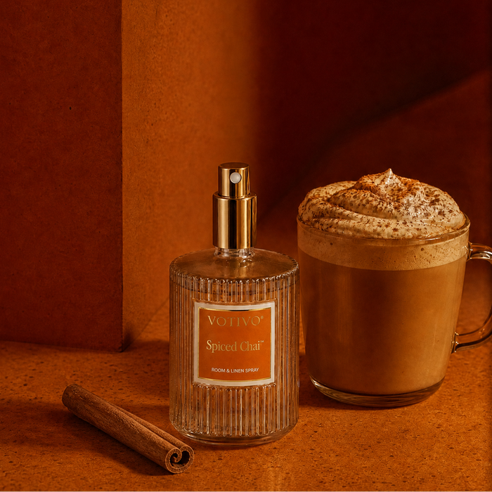 A Spiced Chai room spray bottle with a gold spray top stands next to a frothy latte in a glass mug, with a cinnamon stick on a warm terracotta surface.