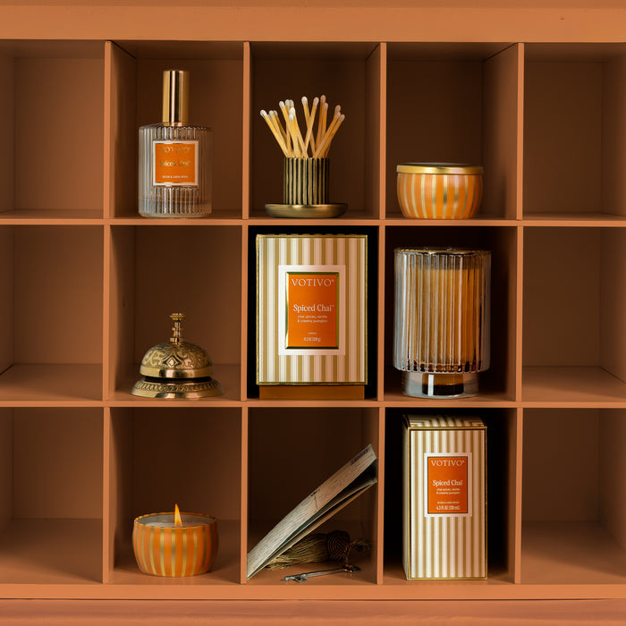 A warm-toned shelving unit with multiple cubbies displaying coordinated spice chai home fragrance and decor items, including reed diffusers, candles, and a boxed scented candle. The products feature striped packaging and orange labels.
