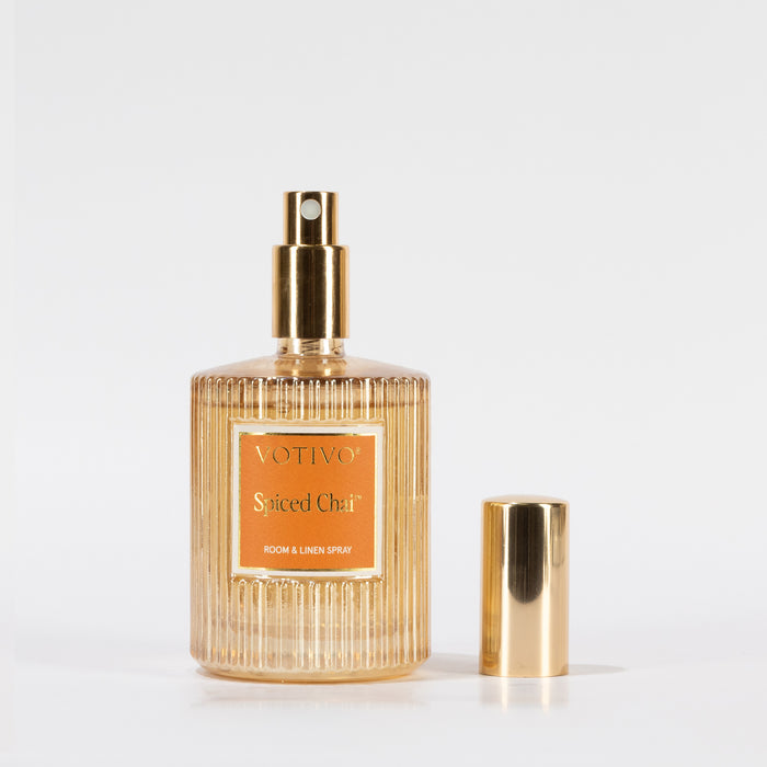 A gold and clear ribbed glass bottle labeled “Votivo Spiced Chai Room & Linen Spray” stands upright with its gold cap beside it on a white background, adding a warm, inviting scent to any room or fabric.