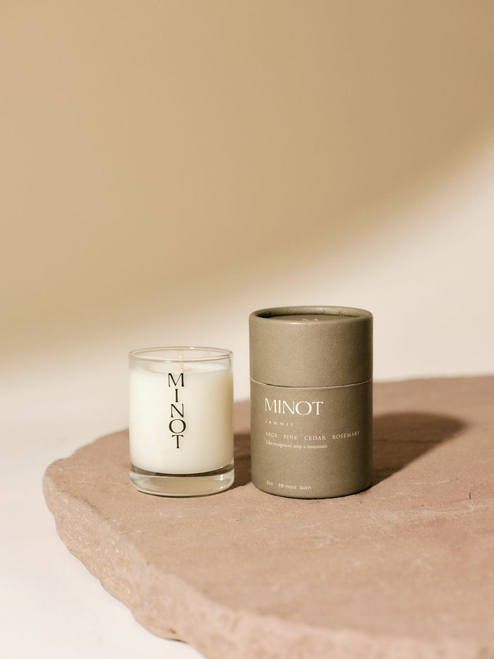 A MINOT Summit mini candle in a glass holder sits beside its cylindrical packaging on a textured stone surface.