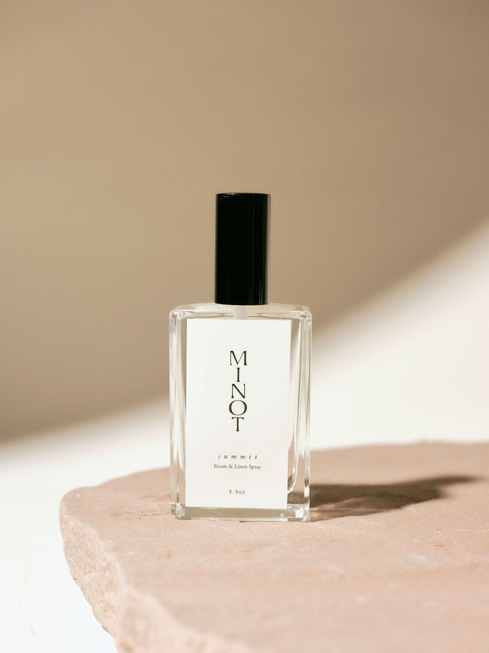 Clear glass bottle of MINOT Summit room and linen spray with black cap, placed on a textured stone surface, highlighted by soft natural light.