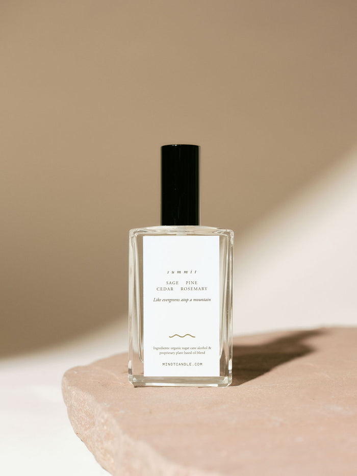 A bottle of MINOT's Summit room and linen spray with sage, pine, cedar, and rosemary scents sits on a stone surface, with a minimalistic design and soft lighting casting gentle shadows.