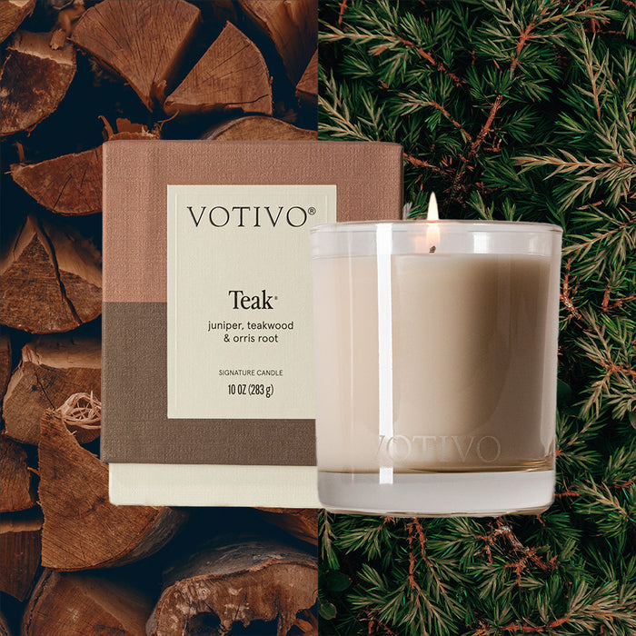 A Votivo Teak 10 oz. Signature Candle rests by its box, framed by stacked firewood and pine branches to emphasize its natural, woody scent and premium soy blend wax.