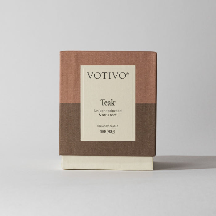 The Votivo Teak 10 oz. Signature Candle features a woody scent of juniper, teakwood, and orris root. Presented in a tan, brown, and cream color-block box, this soy blend wax candle stands out against a plain light background.