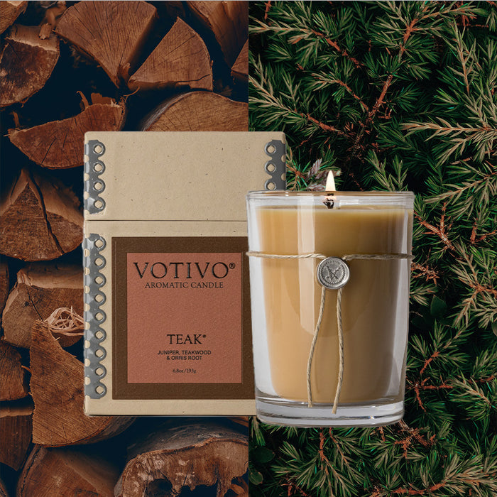 The Votivo Teak 6.8 oz. Aromatic Candle, made from a soy blend wax, stands by its box with stacked wood logs on the left and green pine branches on the right.