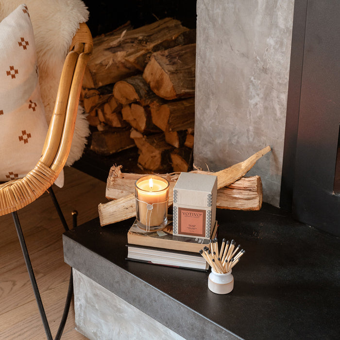 The Votivo Teak 6.8 oz. Aromatic Candle rests on stacked books beside its box, with matches nearby; firewood and part of a cushioned chair appear next to a cozy fireplace in the background.