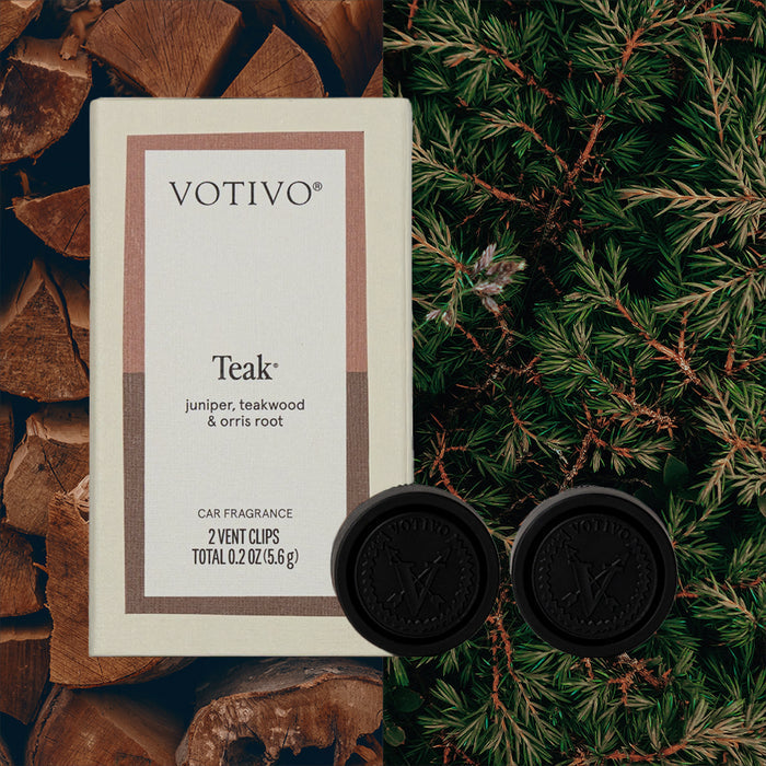 A Votivo Teak Car Fragrance (2 Pack) and two black vent clips are displayed against stacked wood and lush pine branches, evoking the inviting teak aroma.