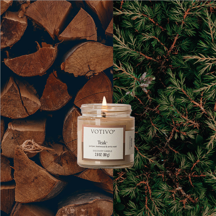 A lit Teak Discovery Candle by Votivo is centered between stacked firewood on the left and green pine branches on the right, evoking natural, woody fragrance notes.