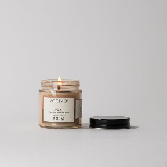 A lit Votivo Teak Discovery Candle with a woody teak fragrance sits in a small glass jar on a white surface, its black lid beside it. The label highlights notes of juniper, tobacco, roots, and soy blend wax.