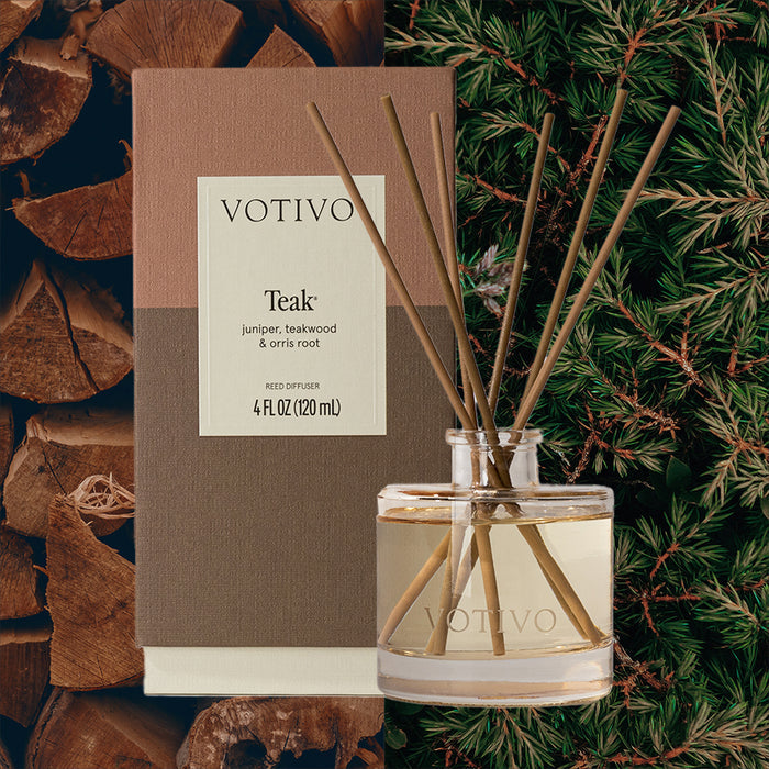 A Votivo Teak Reed Diffuser with fiber reeds and brown packaging stands next to the clear glass vessel of scented liquid. Stacked wooden logs and green pine branches in the background evoke its woody fragrance.