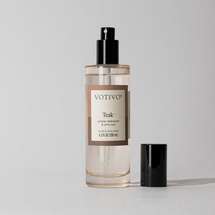 A clear Votivo Teak Room & Linen Spray bottle with a black spray nozzle sits on a light gray background. Its label notes a woody scent blending juniper, teakwood, and orris root.