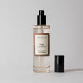 A clear Votivo Teak Room & Linen Spray bottle with a black spray nozzle sits on a light gray background. Its label notes a woody scent blending juniper, teakwood, and orris root.
