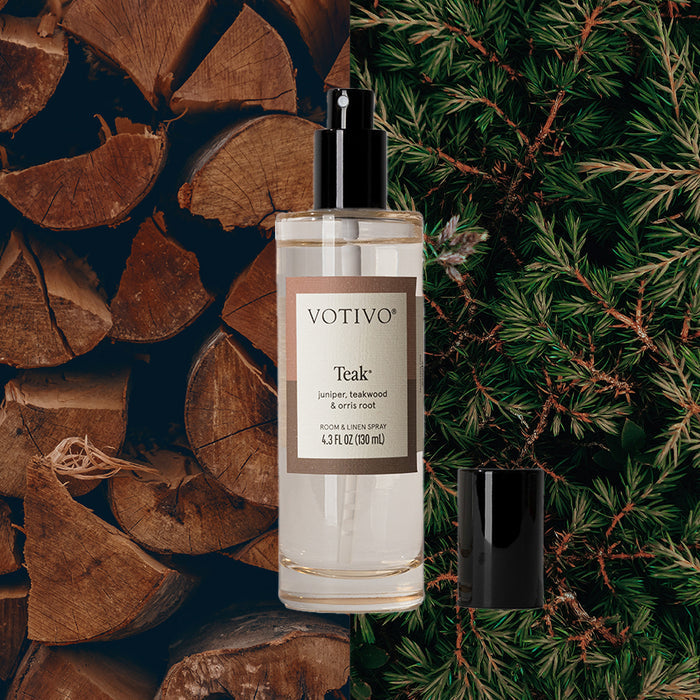 The Votivo Teak Room & Linen Spray, featuring a black cap, is set among stacked wood and pine branches, evoking a natural, woody aroma.