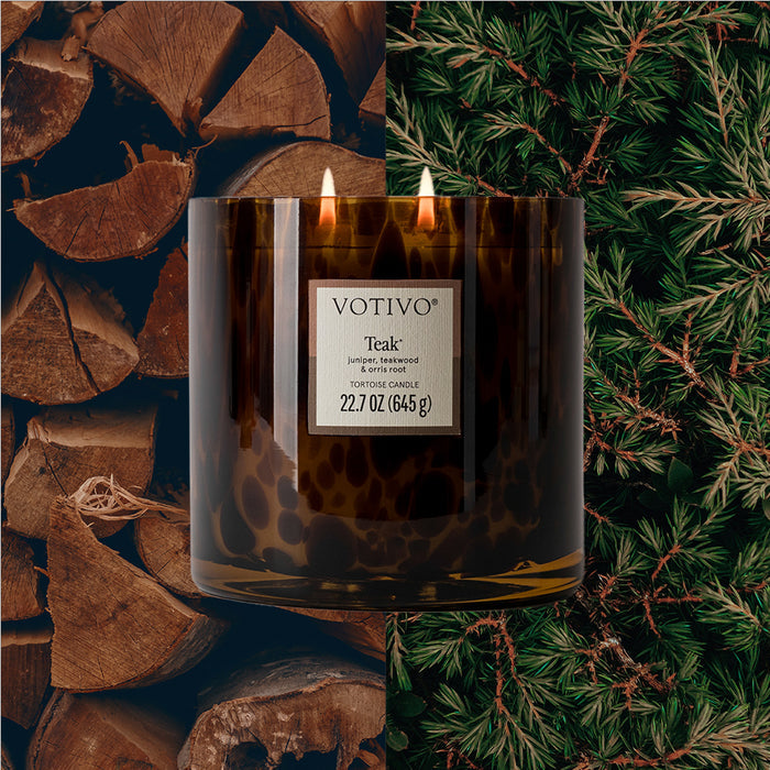 A Votivo Teak Tortoise Candle in brown glass with two flames sits centered over firewood and pine, filling the air with rich teak fragrance from its soy blend wax.