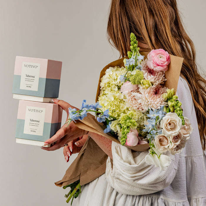 A woman in a white shirt holds a bouquet of pastel flowers in one arm and two Votivo Tuberosa 10 oz. Signature Candles in the other hand against a plain background.