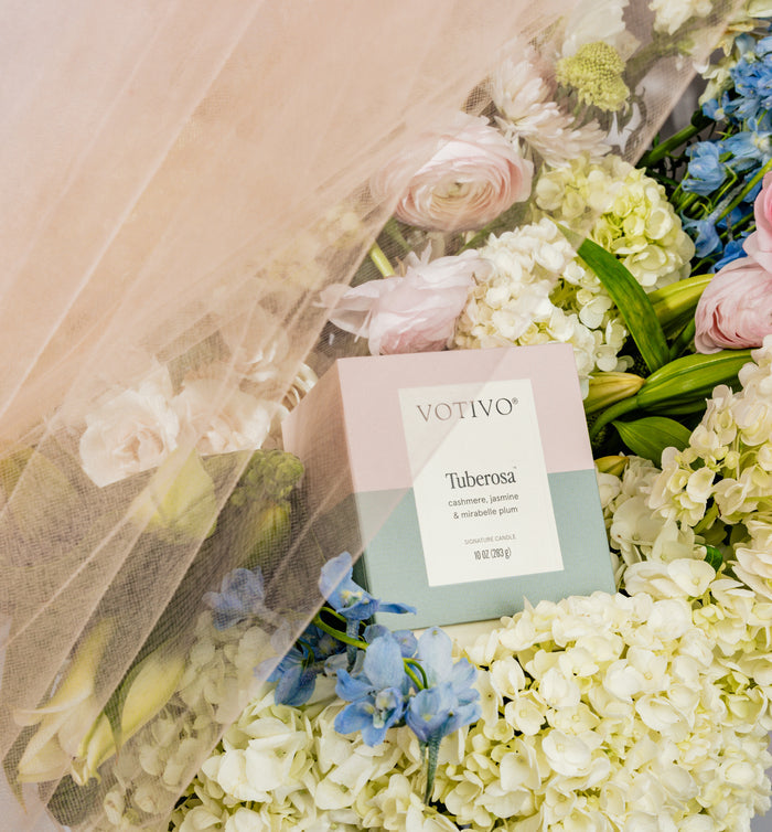 A Votivo Tuberosa 10 oz. Signature Candle rests among white hydrangeas, blue delphiniums, and pink roses, partially draped in sheer peach fabric.